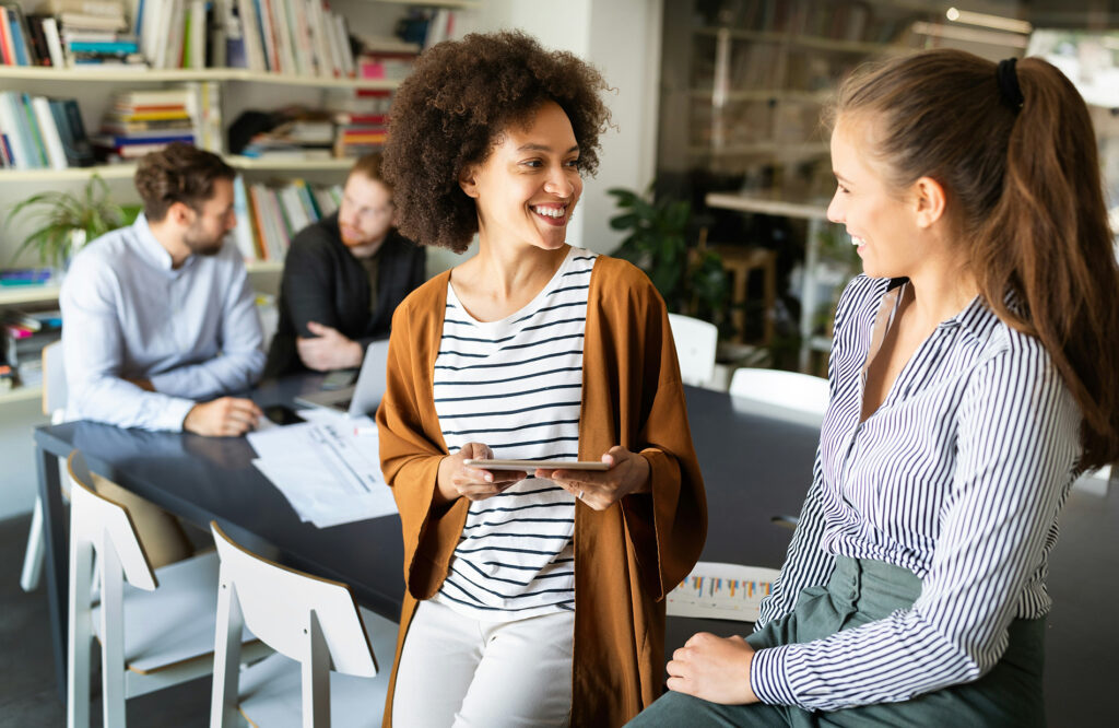 Duas colegas de trabalho sorrindo durante conversa espontânea no escritório, com equipe ao fundo