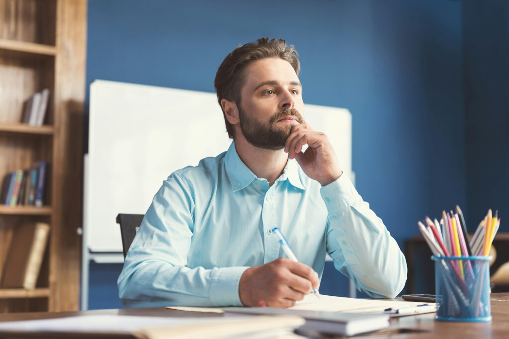 Homem branco de barba, sentado à mesa com caneta na mão, olhando pensativo para o horizonte