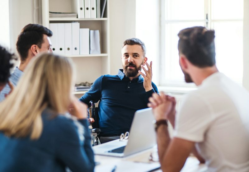 Homem lidera conversa em reunião de equipe, com colegas ao redor prestando atenção em escritório iluminado.