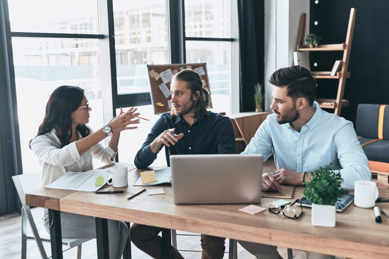 Equipe conversando de forma engajada ao redor de uma mesa de trabalho, com notebook e anotações, em um ambiente de escritório